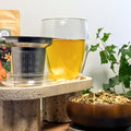 Double-walled Creano 400ml tea glass filled with herbal tea placed beside a stainless-steel infuser and glass lid on a stone stand, with loose-leaf tea and green plant in background.