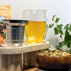 Double-walled Creano 400ml tea glass filled with herbal tea placed beside a stainless-steel infuser and glass lid on a stone stand, with loose-leaf tea and green plant in background.