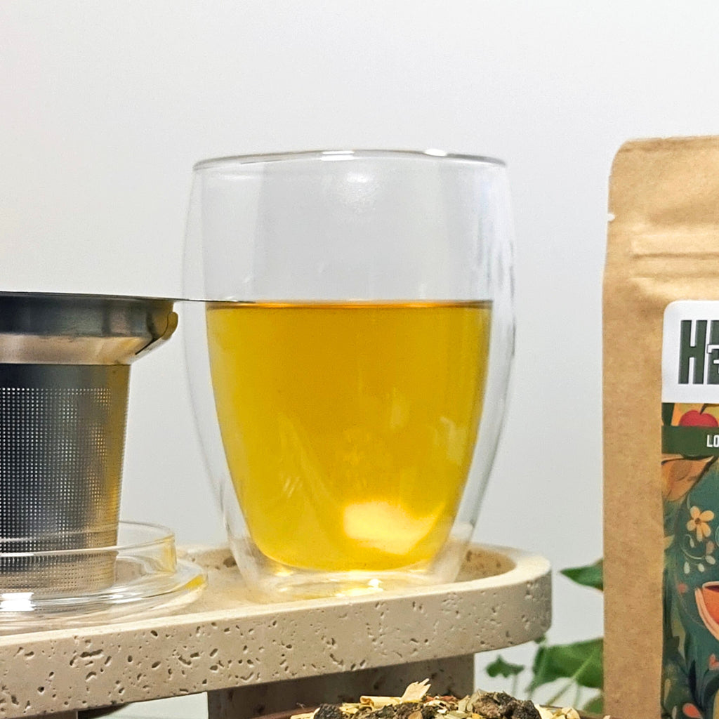 Close-up of the Creano double-walled 400ml tea glass filled with golden herbal tea, next to a stainless-steel infuser on a stone platform.