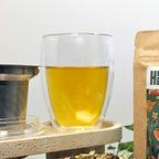 Close-up of the Creano double-walled 400ml tea glass filled with golden herbal tea, next to a stainless-steel infuser on a stone platform.