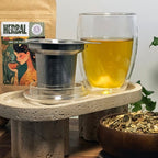 Creano double-walled tea glass with stainless-steel infuser displayed on a textured stone stand, with loose-leaf herbal tea and Holy Tea packaging in the background.