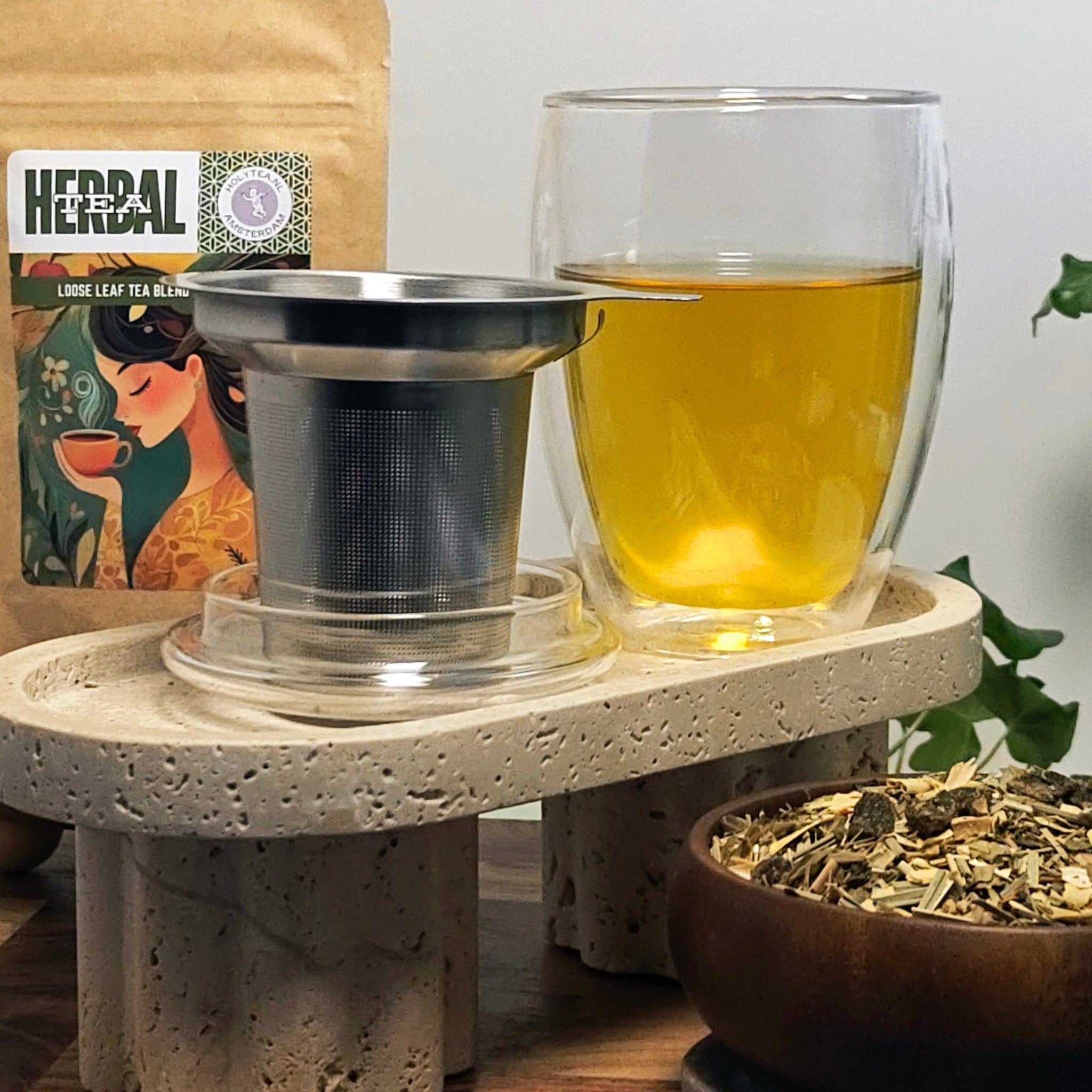 Creano double-walled tea glass with stainless-steel infuser displayed on a textured stone stand, with loose-leaf herbal tea and Holy Tea packaging in the background.