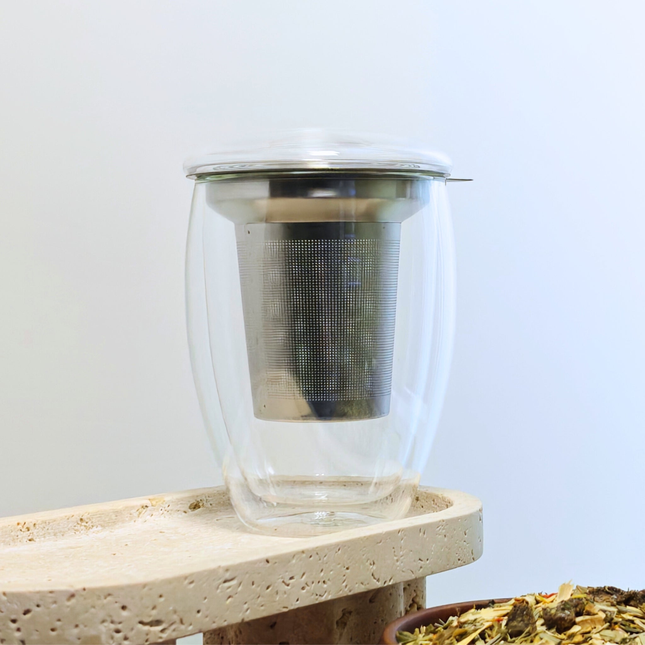 Creano 400ml double-walled glass with stainless-steel tea infuser placed inside, covered with a glass lid, shown on a stone platform with loose herbal tea nearby.