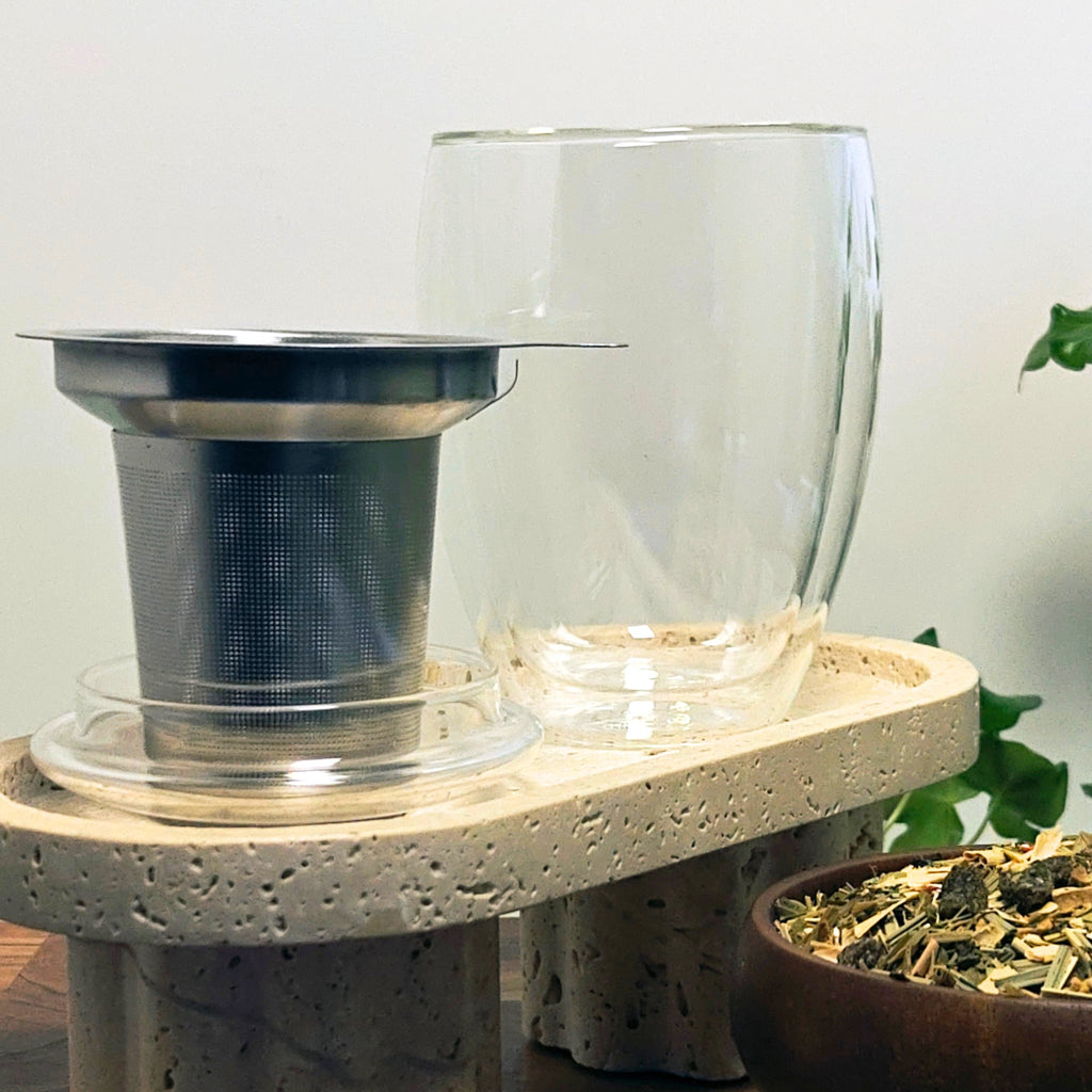 Double-walled Creano tea glass and stainless-steel infuser displayed side by side on a stone stand, with loose-leaf herbal tea in a wooden bowl in the foreground.