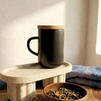 Minimal black stoneware Juliet mug with closed acacia wood lid for heat retention sitting on a terrazzo stand.