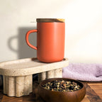 Paprika red stoneware Juliet mug with closed acacia wood lid for heat retention sitting on a terrazzo stand.