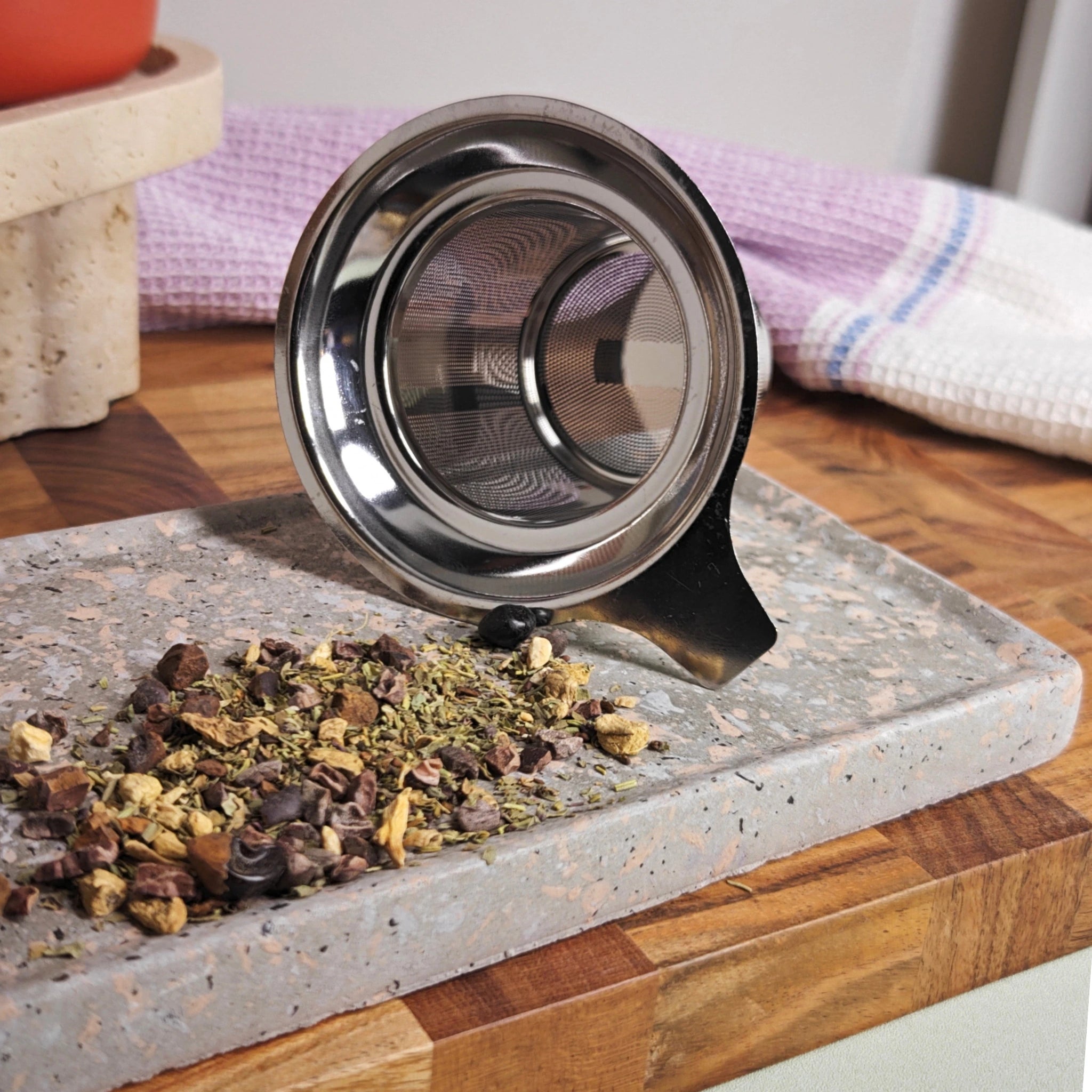 Detail of fine mesh stainless steel infuser for loose-leaf tea with dried herbs and Paprika mug in background.