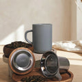 Juliet Perle grey stoneware tea mug 475ml with stainless steel infuser and acacia lid next to loose-leaf tea.