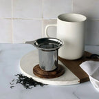 Juliet tea filter resting on its acacia wood lid next to a white mug and loose-leaf tea